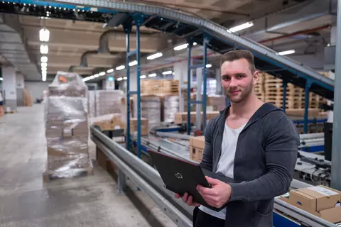 Ein junger Mann lehnt an einem Band und hält einen Laptop  